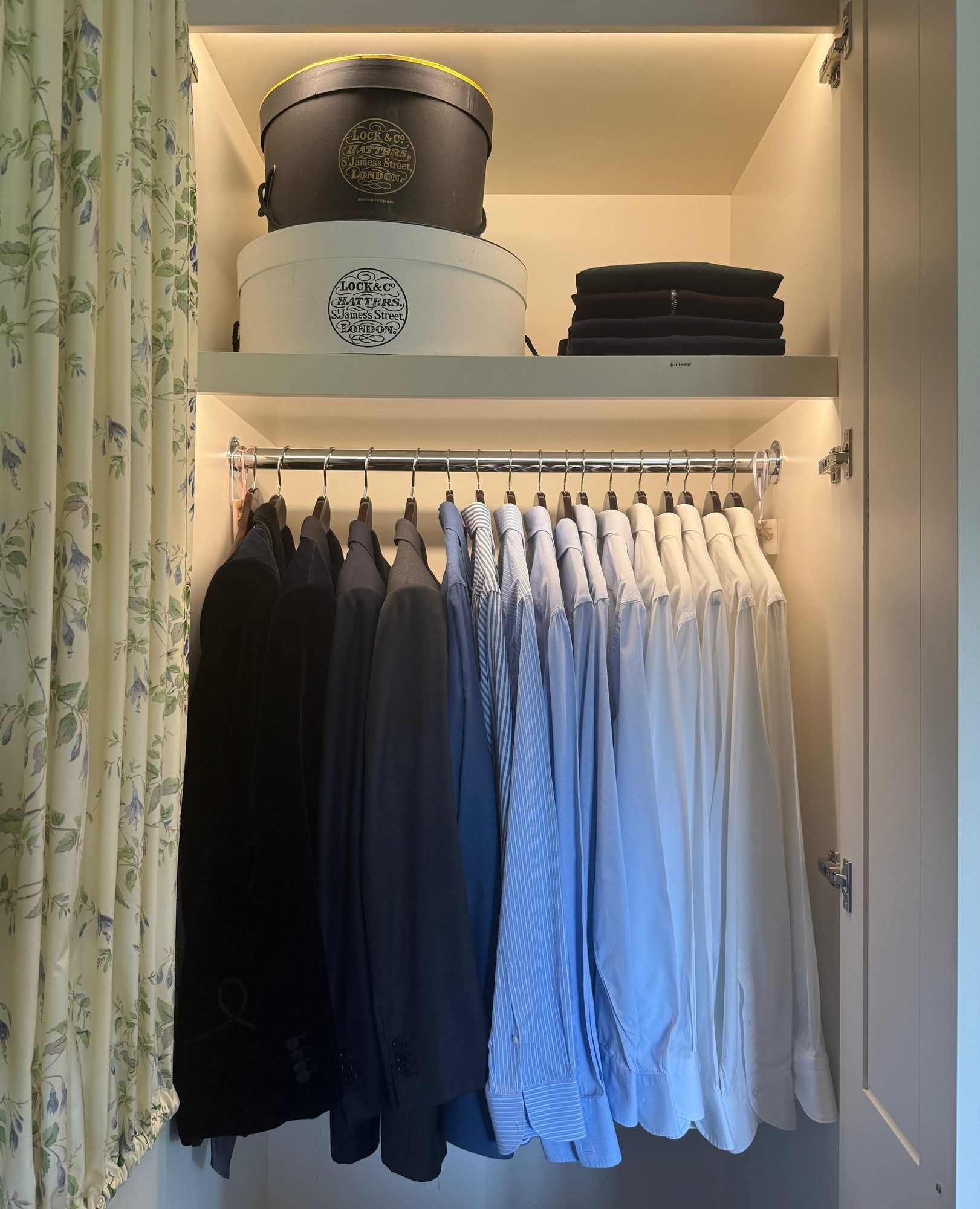 Shirts hung by colour beneath hatboxes on a wardrobe shelf.
