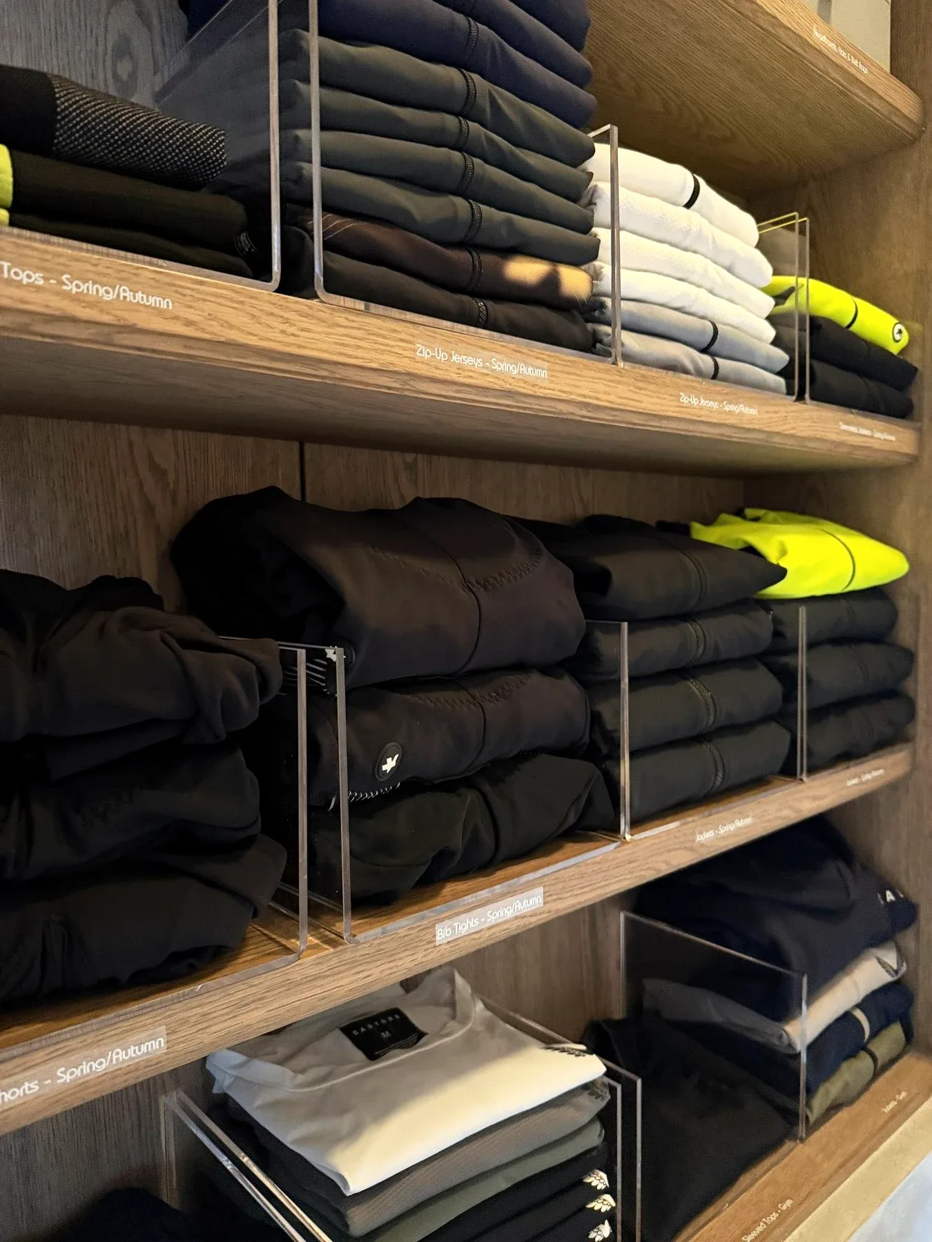 Folded sportswear in clear dividers on open timber shelves, each shelf edge labelled by kind and season.