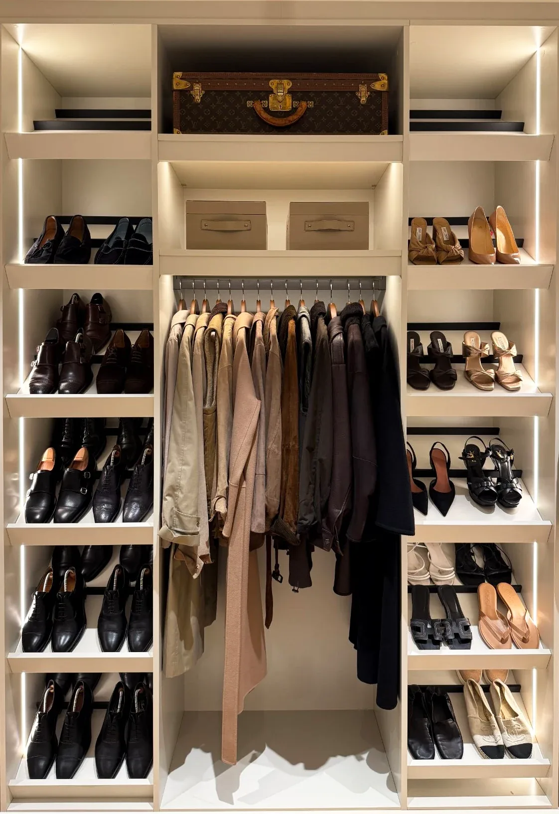 A lit dressing bay with a central rail of coats, shoe shelves either side and a trunk above.