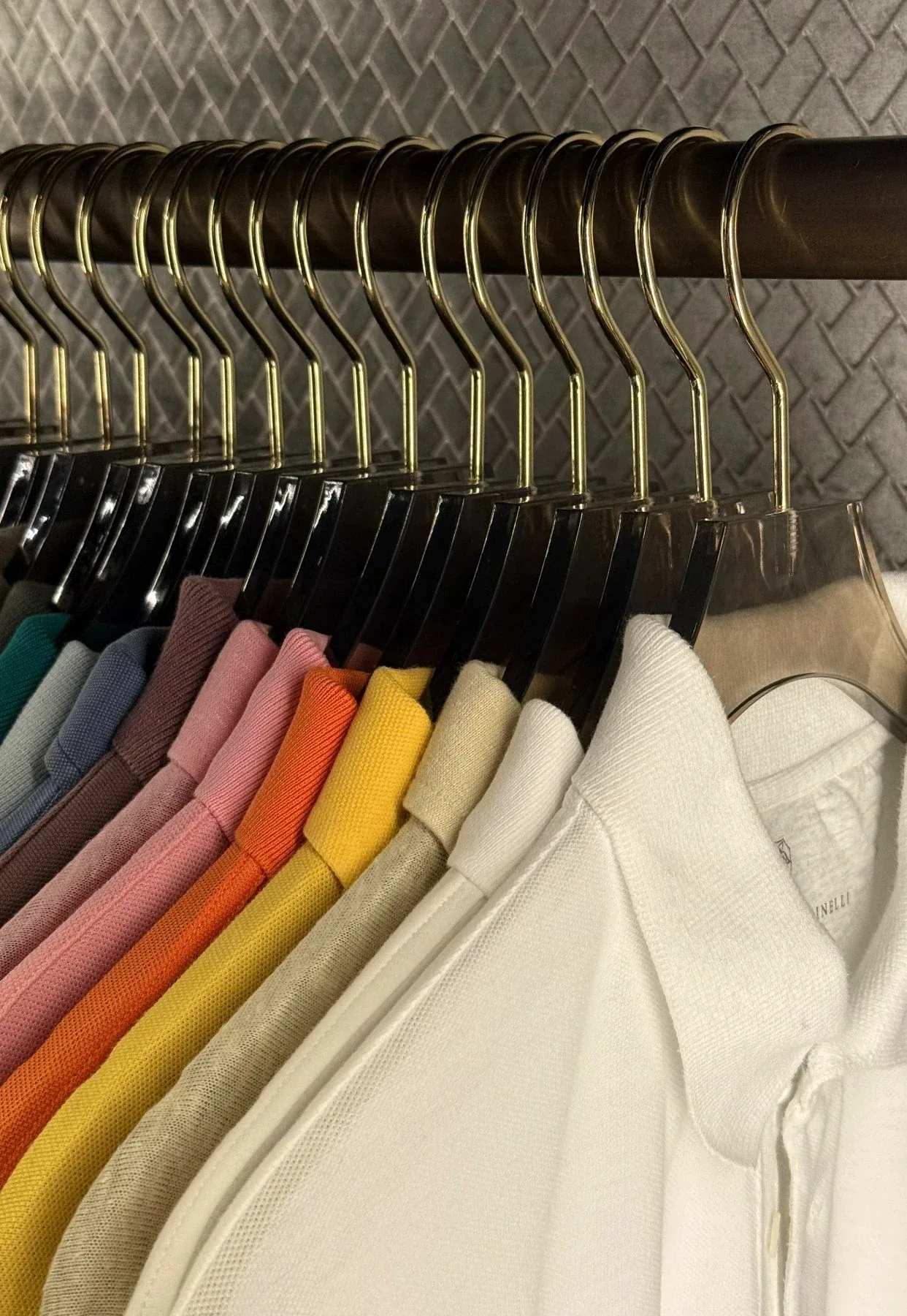 Polo shirts on matching hangers, graded from white through yellow and pink to blue, against a herringbone wall.