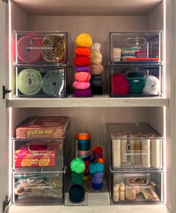 A lit craft cupboard of labelled clear drawers holding paper rolls, yarn and wooden pieces.