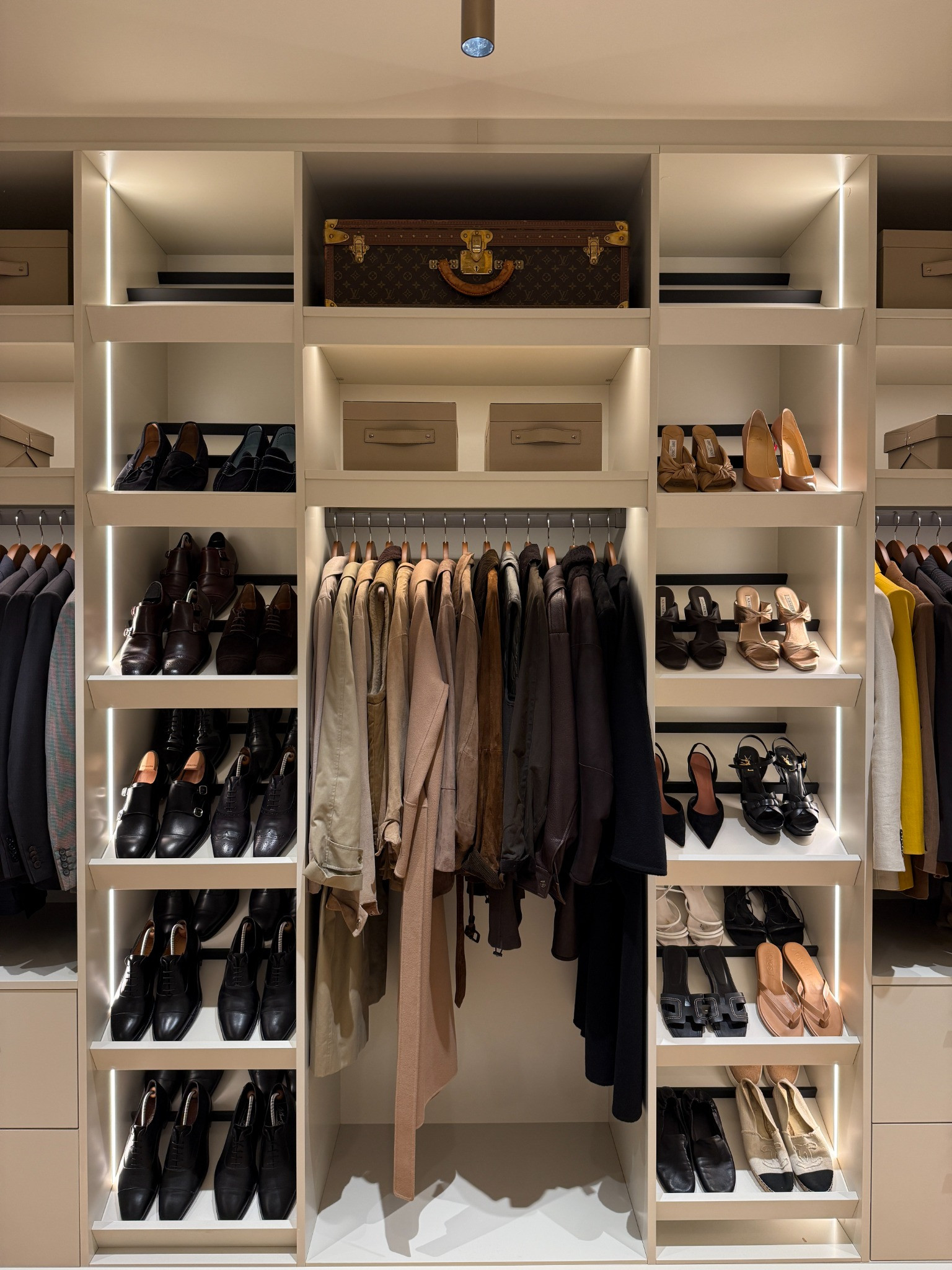 A walk-in wardrobe with lit shelves of shoes and boxes either side of a rail of coats on matching wooden hangers.