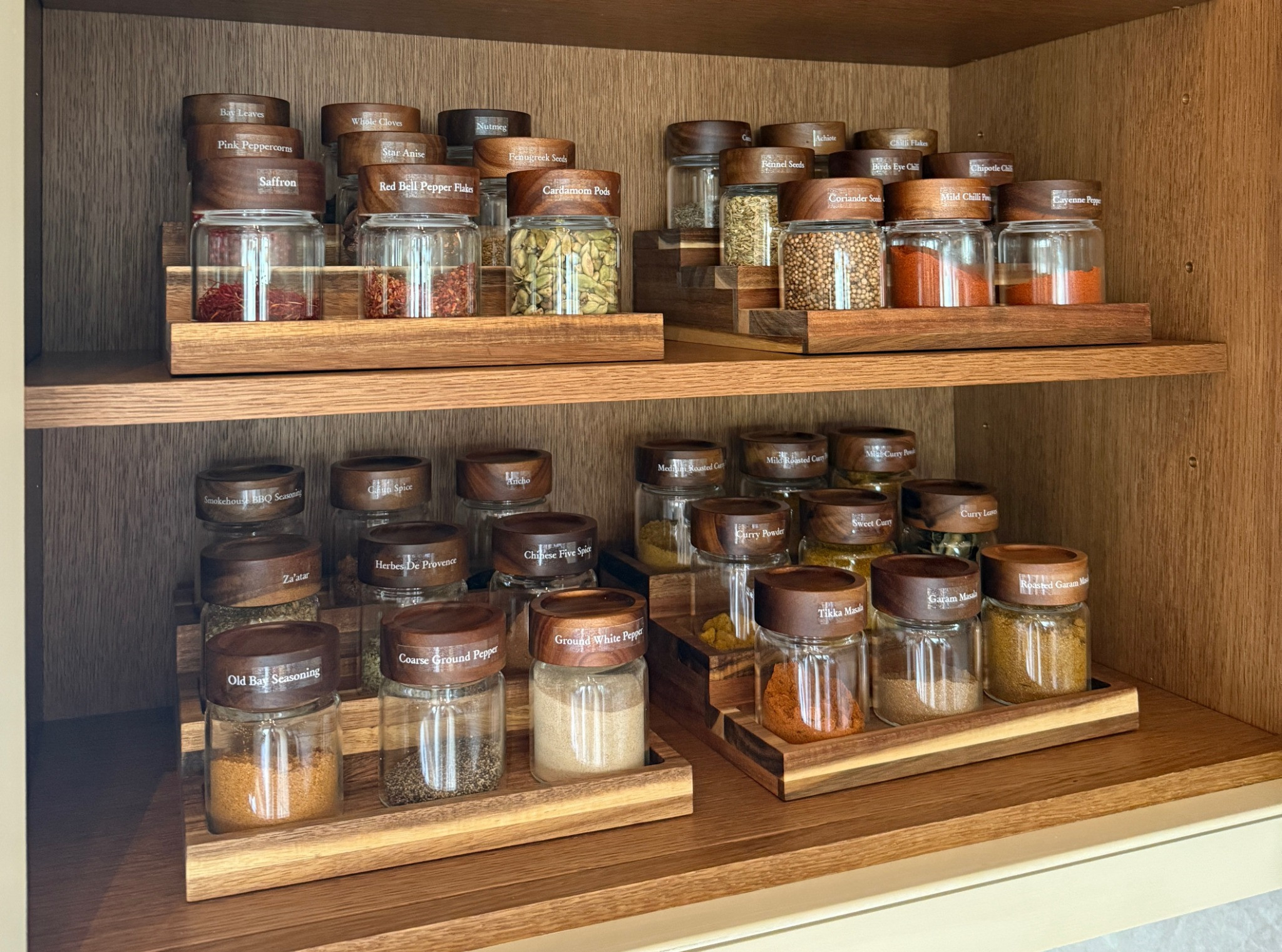 Labelled spice jars with walnut lids on tiered wooden risers in an oak cupboard.
