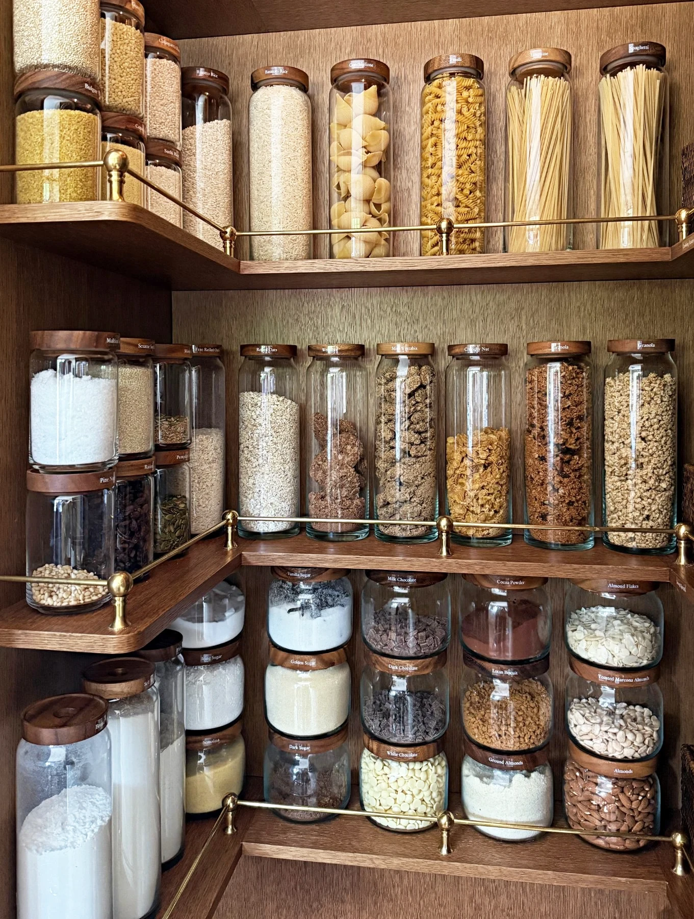 A pantry corner of decanted grains and pasta in tall glass jars on brass-railed shelves.
